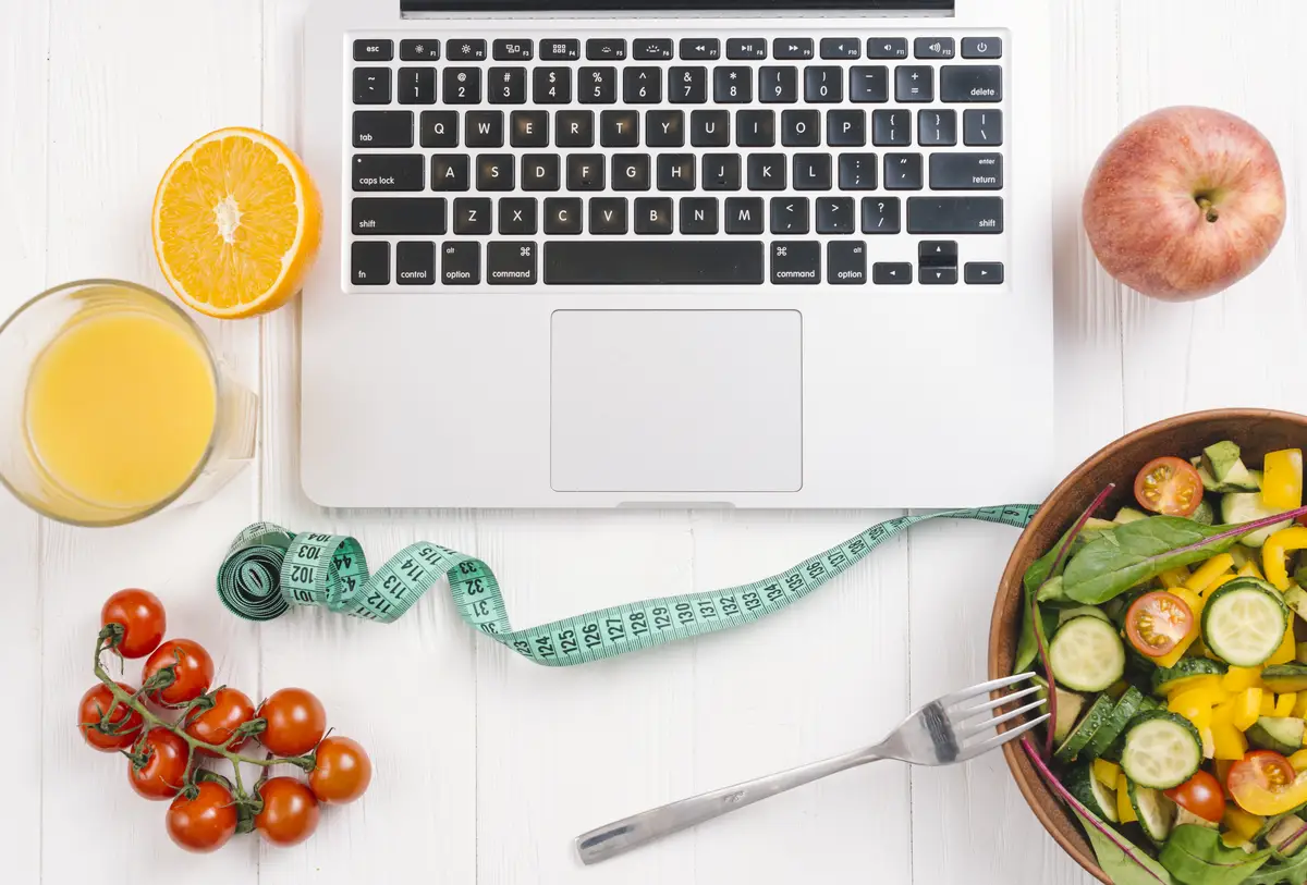 An overhead view of laptop with fresh salad; fruits; juice and cherry tomatoes on white wooden desk