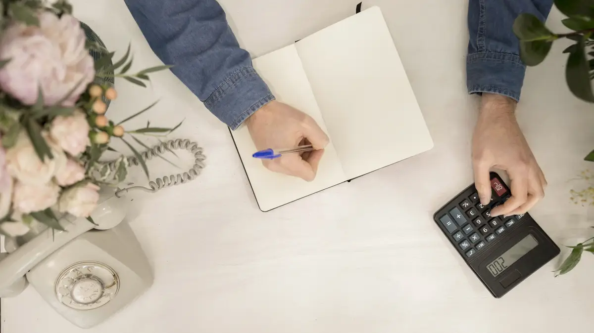 An overhead view of florist writing on notebook using calculator on white desk