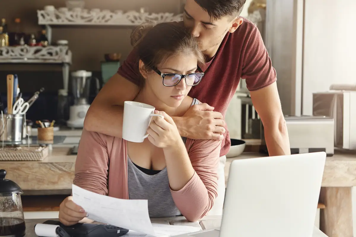 Young couple having credit problem in bank. supportive man hugging and kissing his unhappy wife on her head while she is sitting at kitchen table in front of laptop