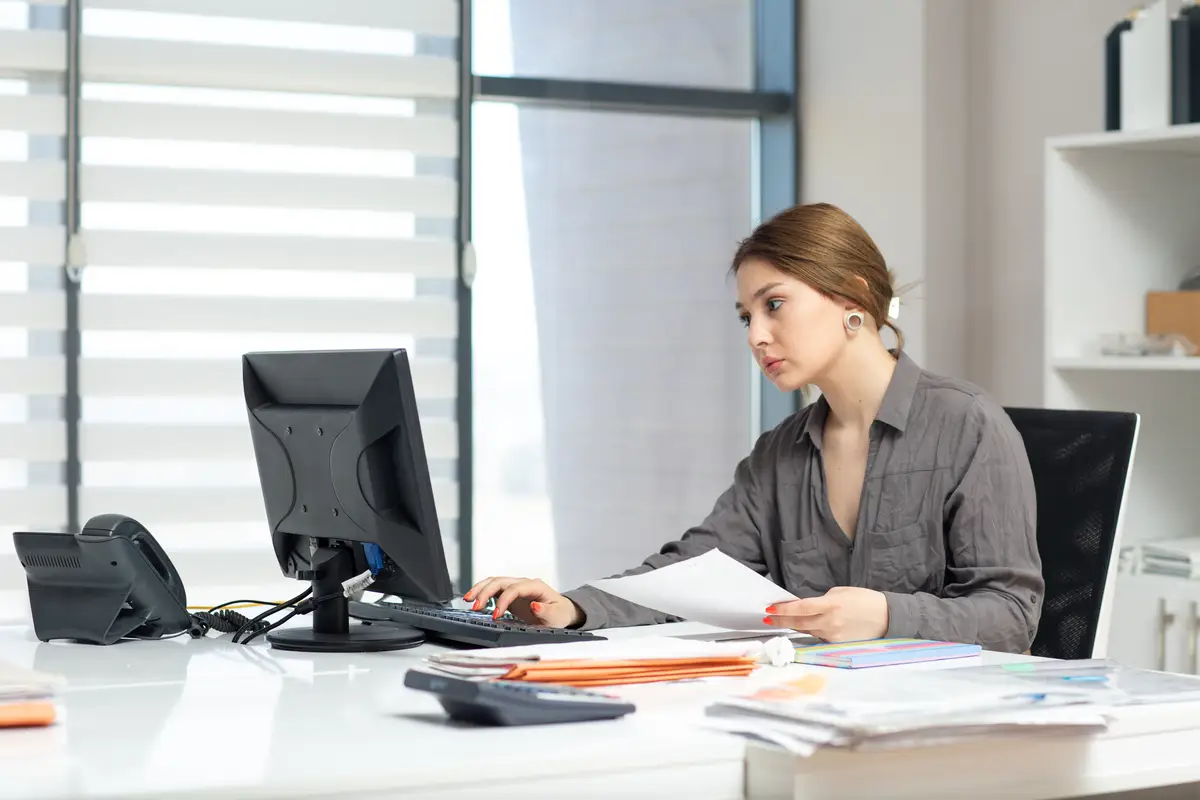 A front view young beautiful lady in grey shirt working on her pc and looking through documents sitting inside her office during daytime building job activity