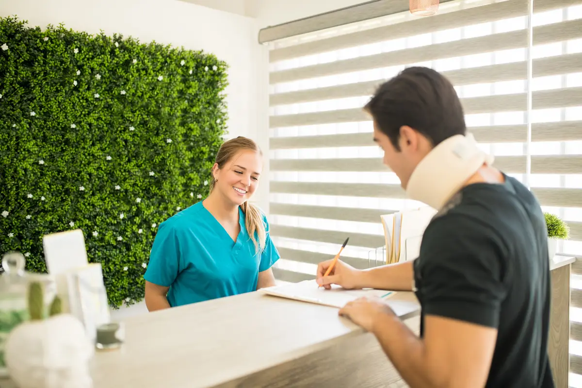 Physical therapist looking at male athlete with cervical collar filling medical form at reception counter in hospital