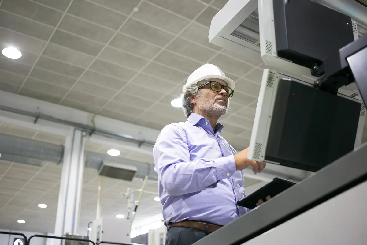 Focused male plant engineer in hardhat and glasses operating industrial machine, pushing buttons on control panel