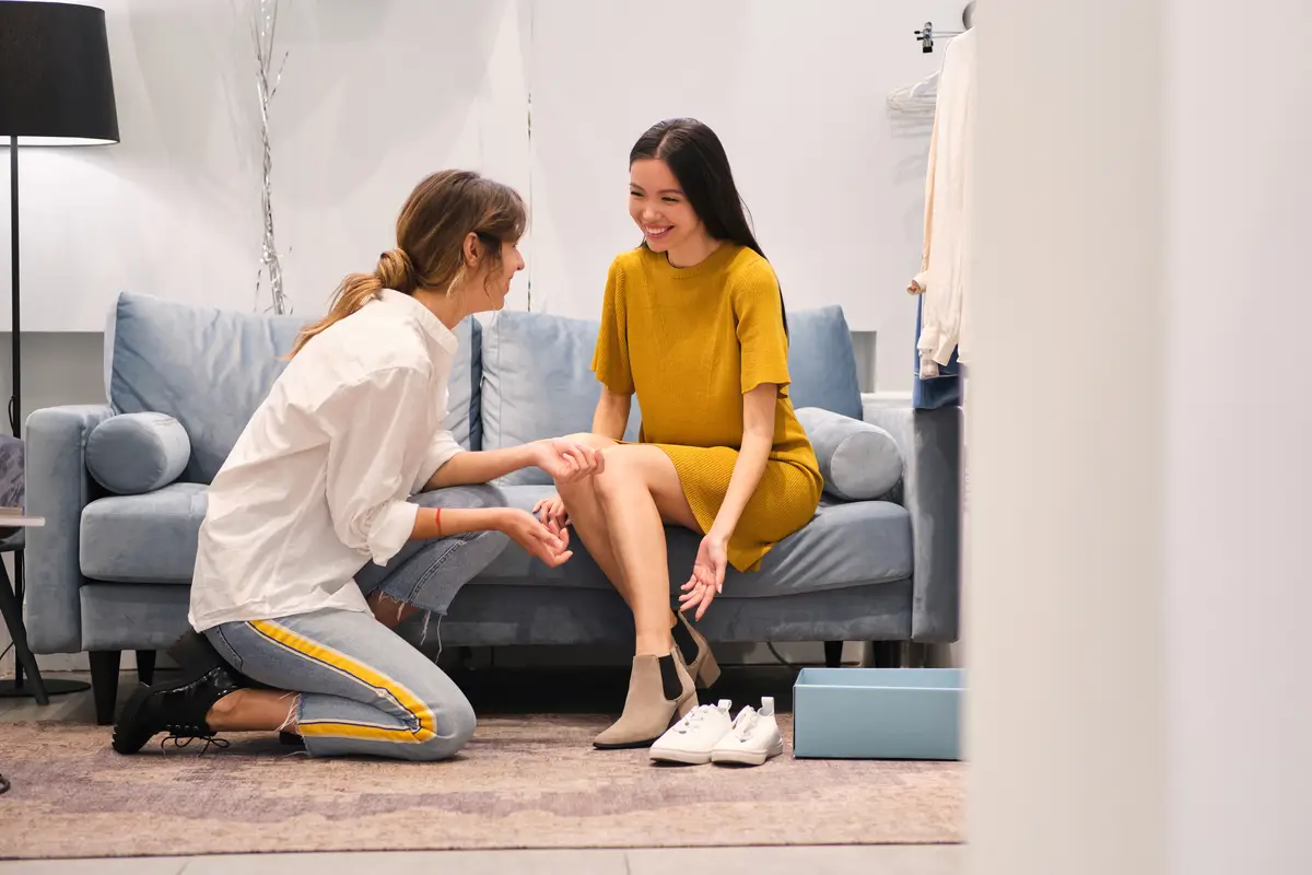 Young fashion consultant joyfully helping trying on boots to cheerful Asian girl in modern showroom