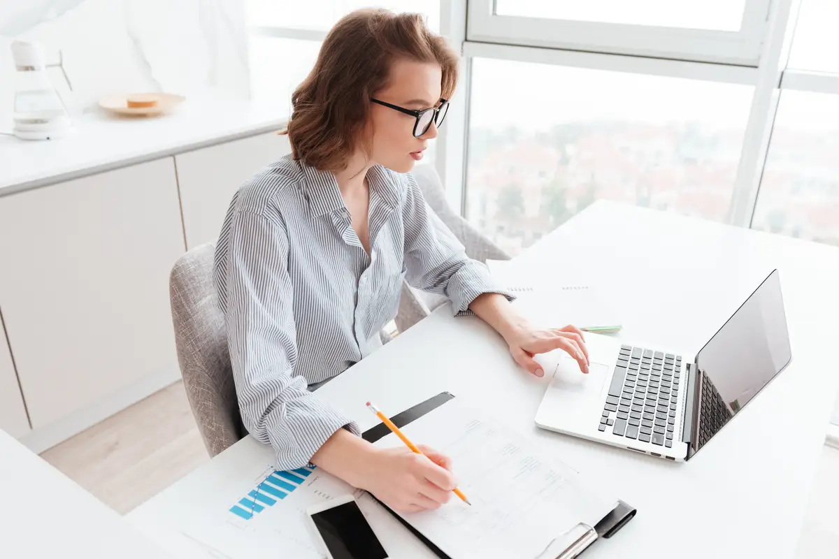 Como manter a contabilidade remota nos EUA: ferramentas úteis 2 charming caucasian businesswoman in striped shirt working with laptop and papers in light apartment