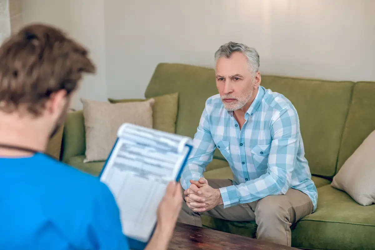 Sad serious mature man dressed in a plaid shirt sitting on the sofa before a dark-haired doctor