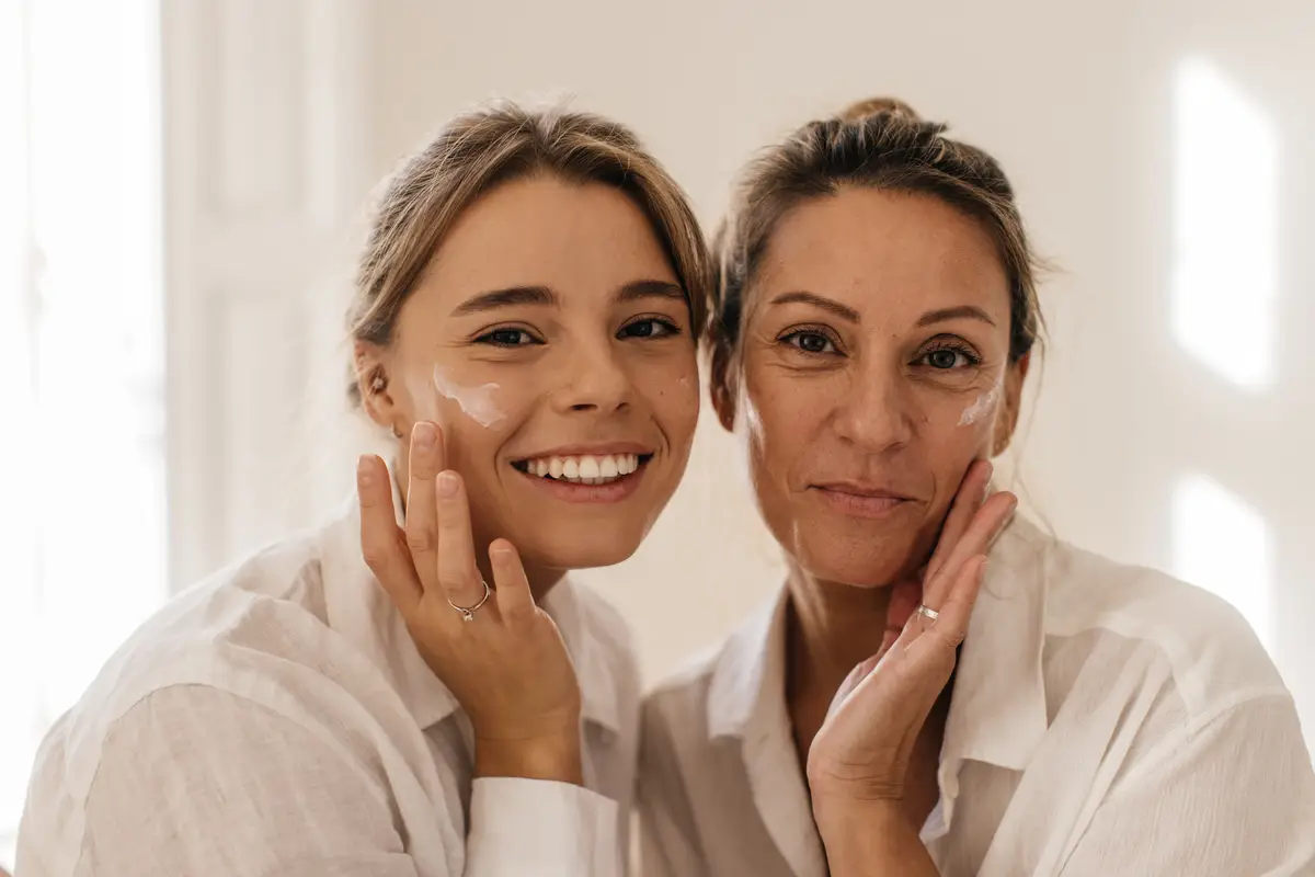 Close-up of two cute caucasian women smearing their face with cream and looking at camera on white background. Beauty and youth concept