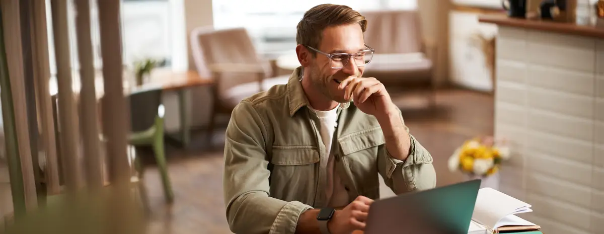 Image of young digital nomad man in glasses sits in cafe works from coffee shop uses laptop in coworking space wears glasses drinks his beverage