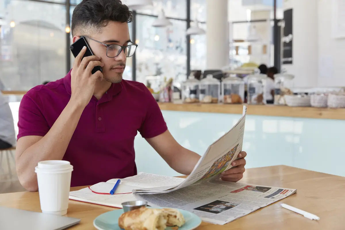 Sideways shot of concentrated entrepreneur reads business newspaper, makes phone call, works remotely from coffee shop, talks on cellular, drinks takeaway coffee, eats dessert. Mass media concept