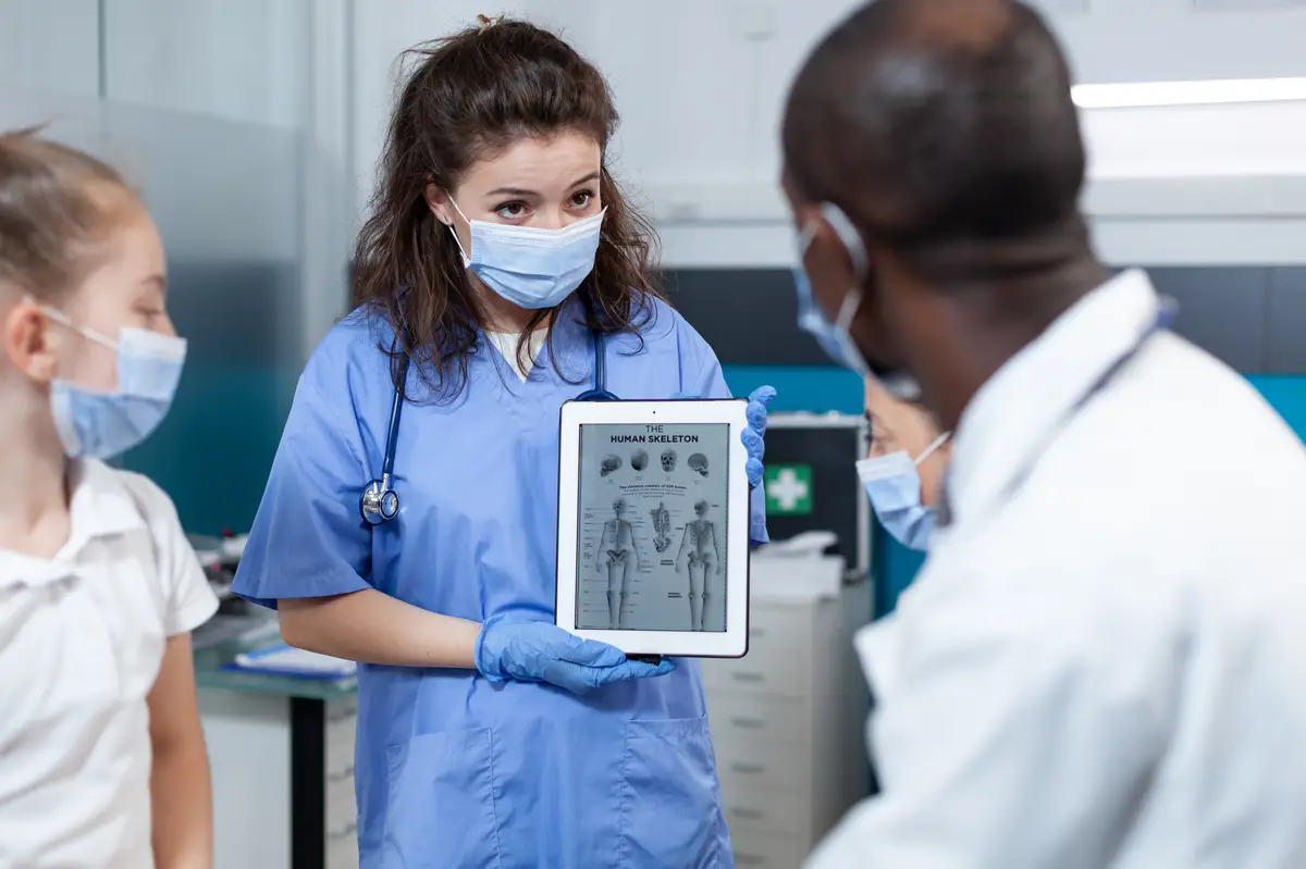 Pediatrician nurse with protective face mask against covid19 holding tablet computer with bones radiography on screen. African american doctor explaining medical expertise in hospital office