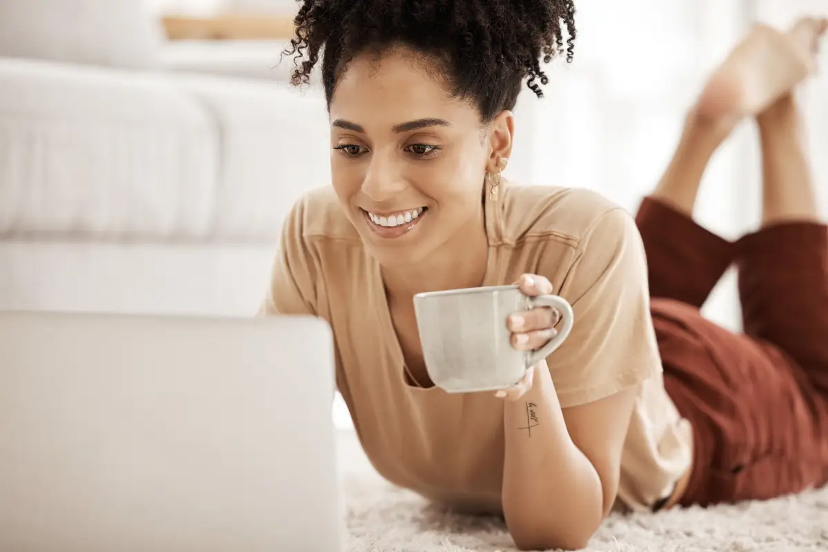 Black woman coffee and home computer of a person watching a video or writing a remote work email Digital employee in the morning on a laptop online shopping or streaming series on a house carpet