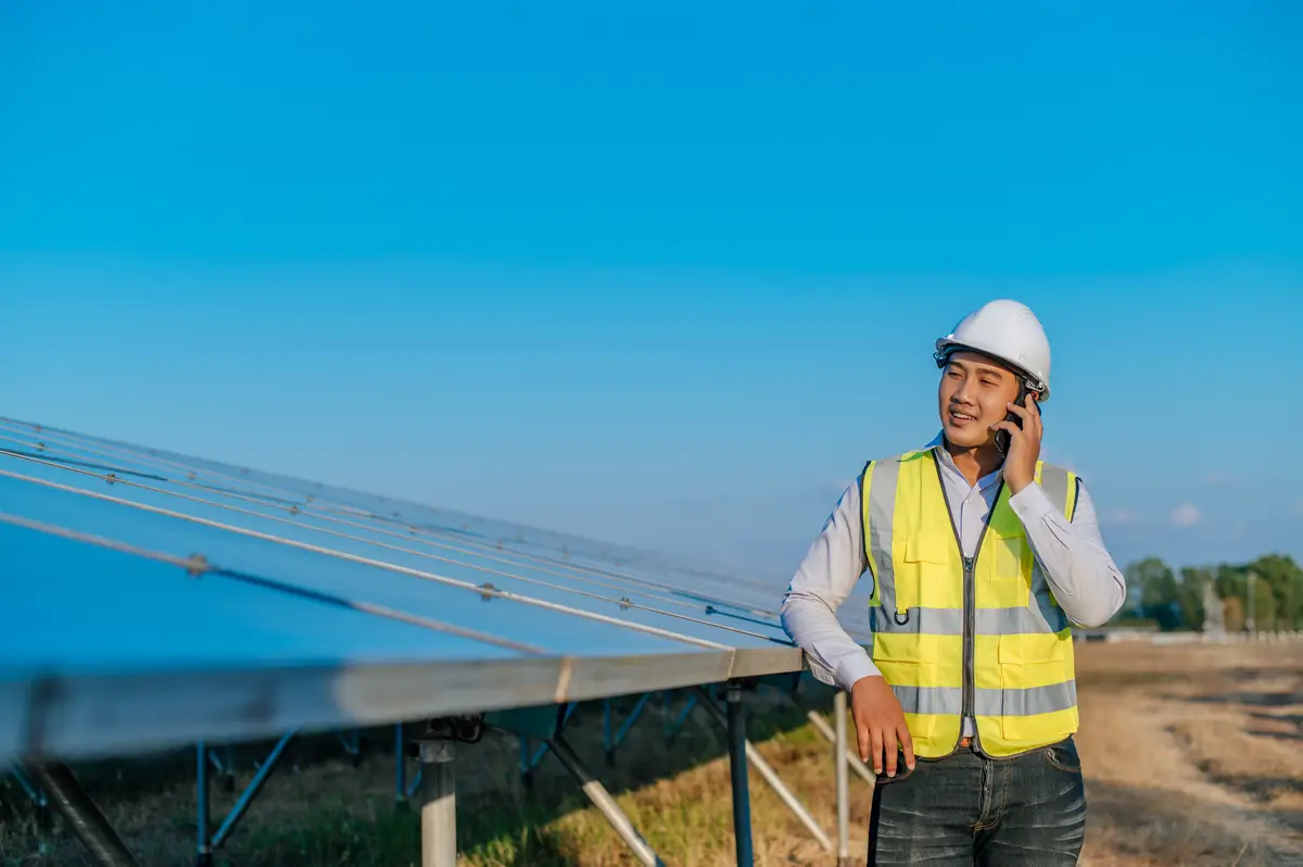 Young Asian technician man standing and talking on smartphone between long rows of photovoltaic solar panels copy space