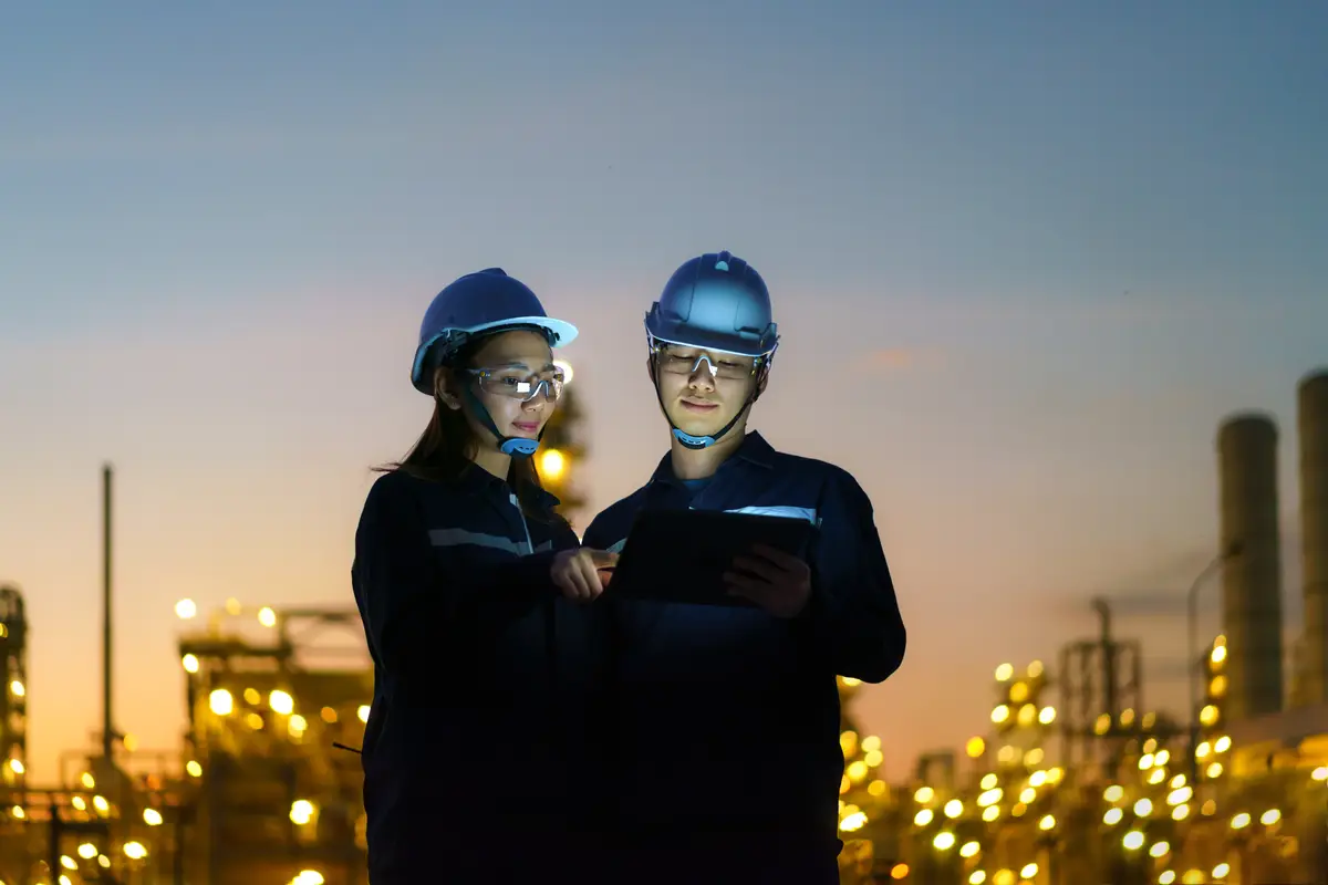 Asian engineers, man and woman are checking the maintenance of the oil refinery factory at night via digital tablets.