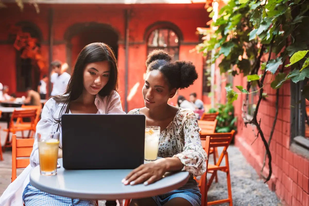 Two beautiful girls thoughtfully using laptop while spending time with cocktails together in cozy courtyard of cafe