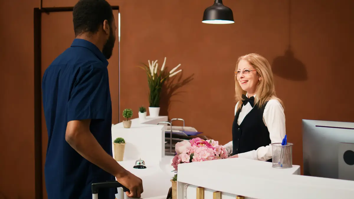 Hotel guest entering lobby with luggage, arriving at tropical summer resort on vacation trip. Male tourist checking in at exotic hotel, talking to front desk staff. Handheld shot.