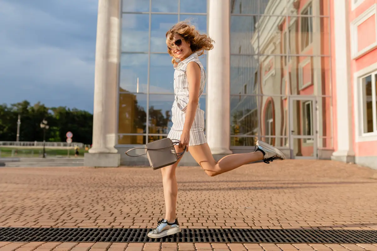 Young attractive stylish woman running jumping funny in sneakers in city street in summer fashion style white dress wearing sunglasses and handbag