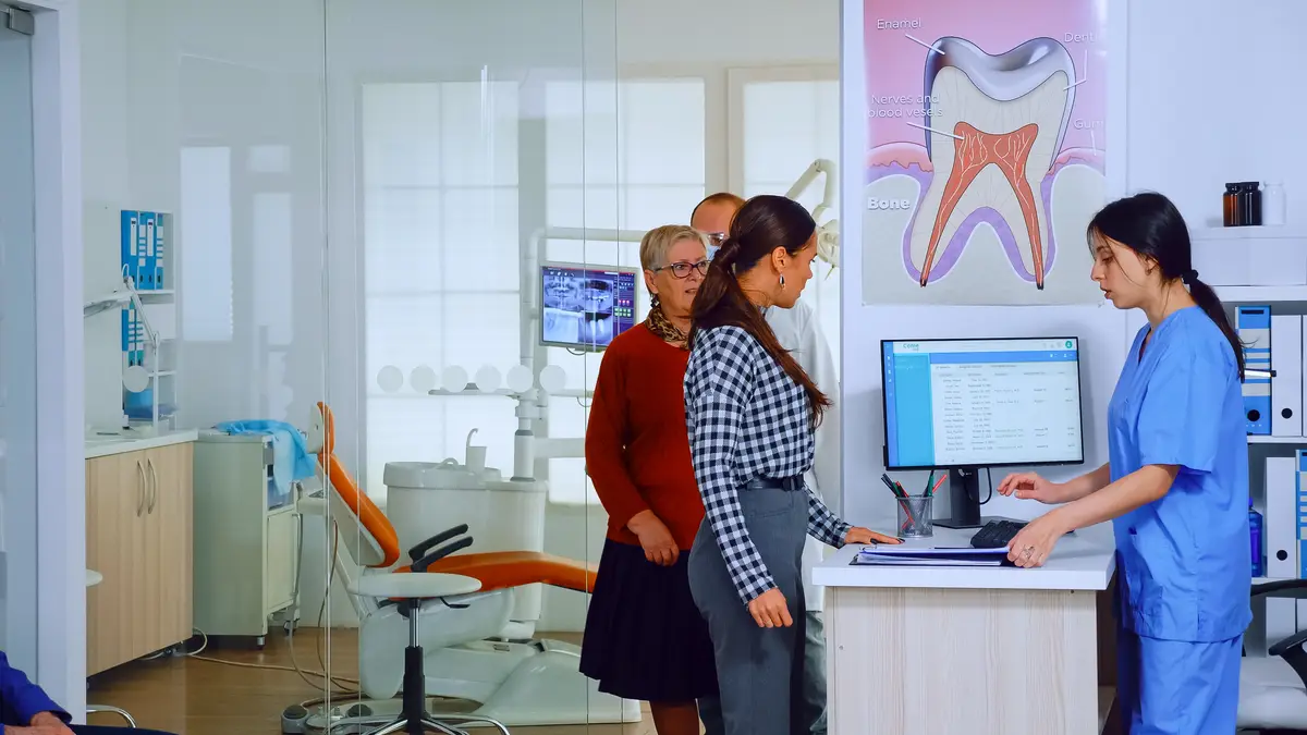 Dentist inviting senior man in consultation dental room while nurse giving to patient form to fill indicating to sit on chair in waiting area. Slow motion shot crowded professional orthodontist office