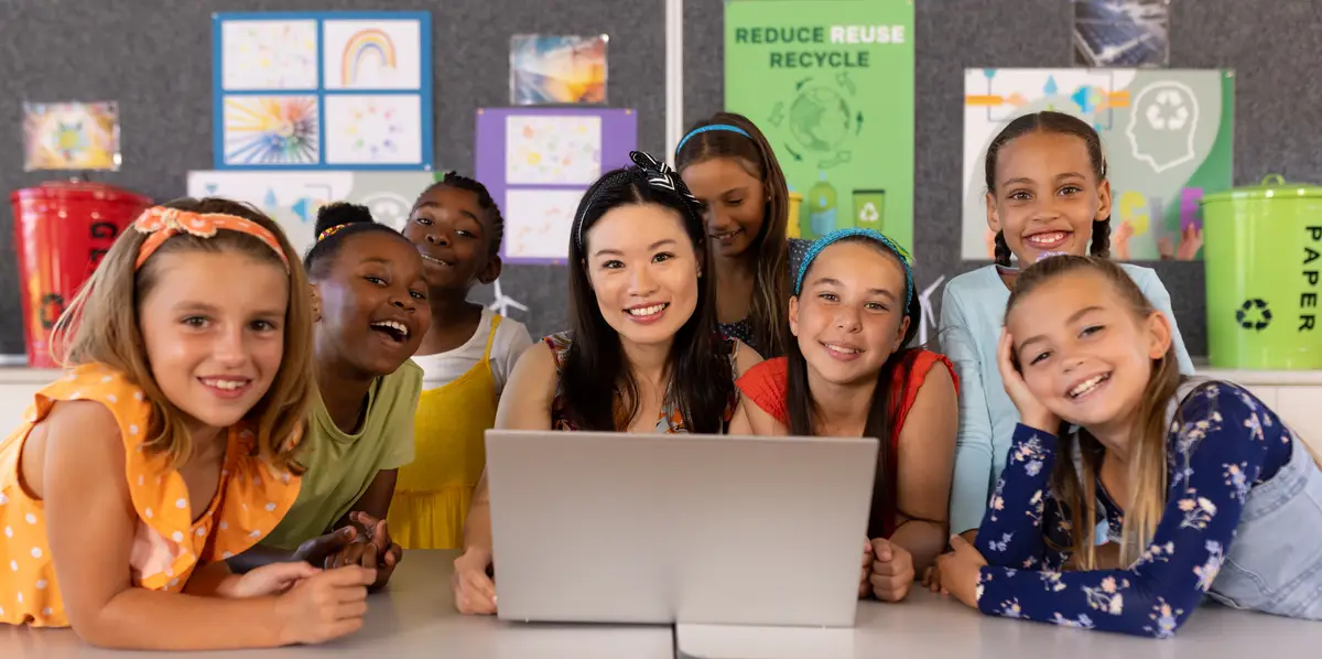 Portrait of smiling diverse female teacher and schoolgirls with laptop on desk in classroom