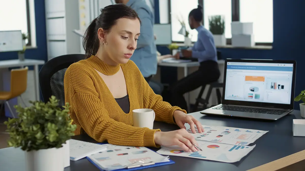 Portrait of startup employee comparing two sheets of paper with charts analyzing financial data while drinking morning coffee. Woman working in busy marketing department sitting at desk with laptop.