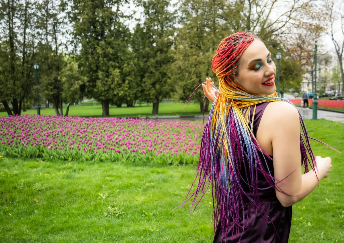 Cute girl with colorful glitter makeup and long multi-colored fake braids in a satin purple dress. Smiles and hugs herself and bright braids on a walk in a blooming spring park.
