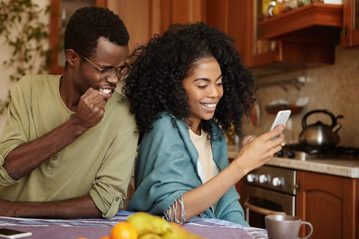 Furious and jealous African American man clenching fist in anger and fury while catching his cheating girlfriend as she messaging her lover on mobile phone having happy and cheerful expression
