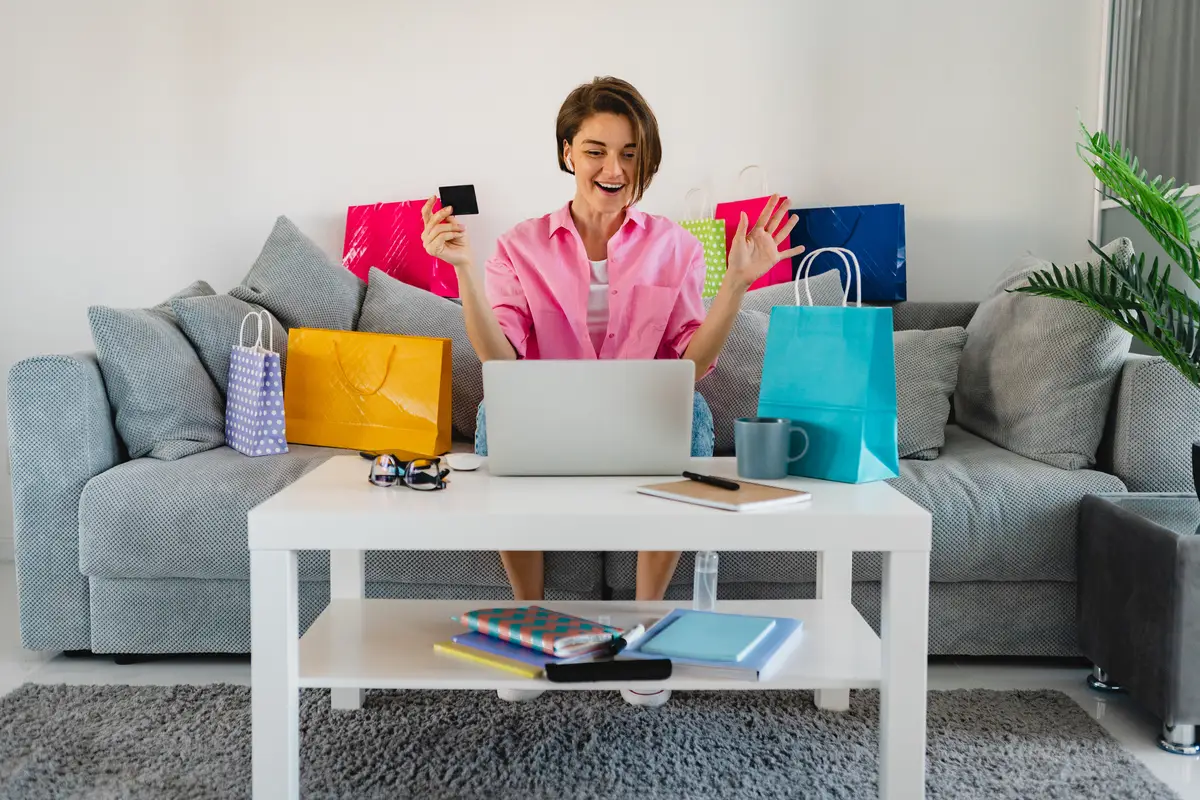 Happy smiling woman in pink shirt on sofa at home among colorful shopping bags holding credit card paying online on laptop