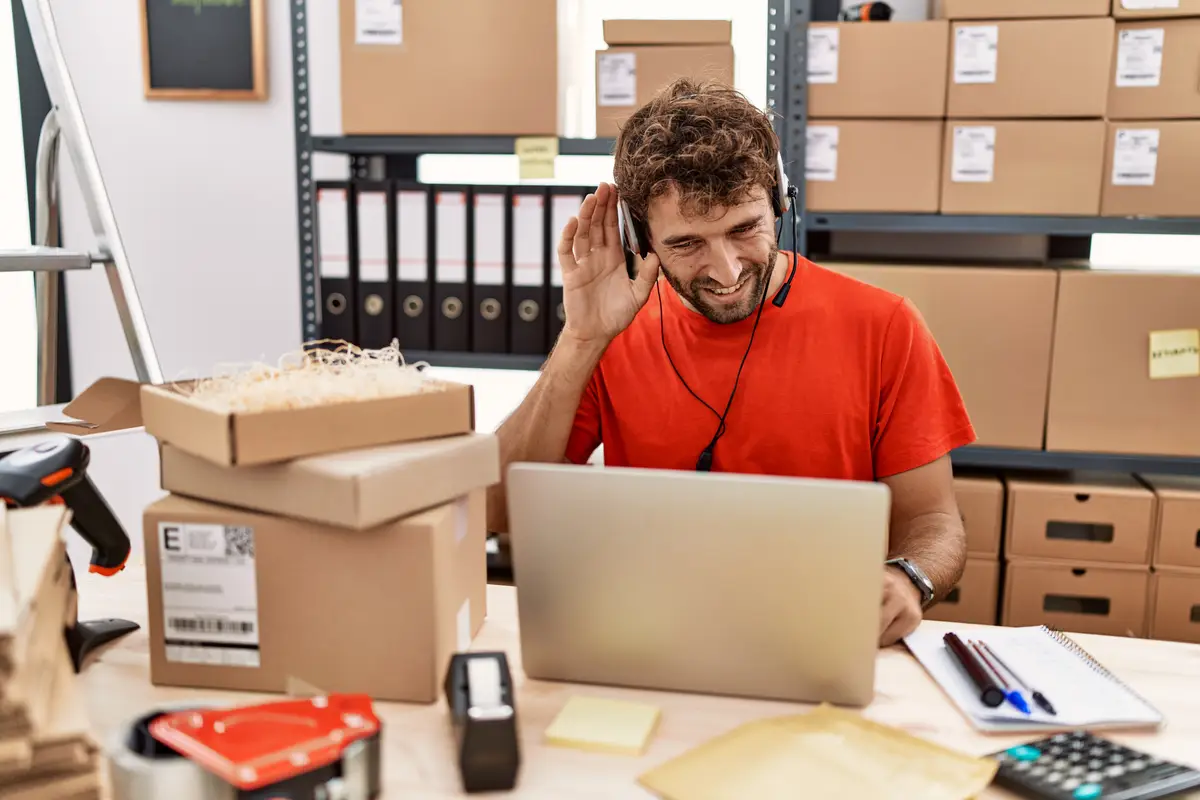 Young hispanic call center agent man working at warehouse smiling with hand over ear listening an hearing to rumor or gossip. deafness concept.