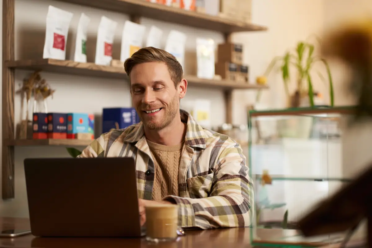 Portrait of cafe owner young man sitting in coffee shop with laptop working on data project looking