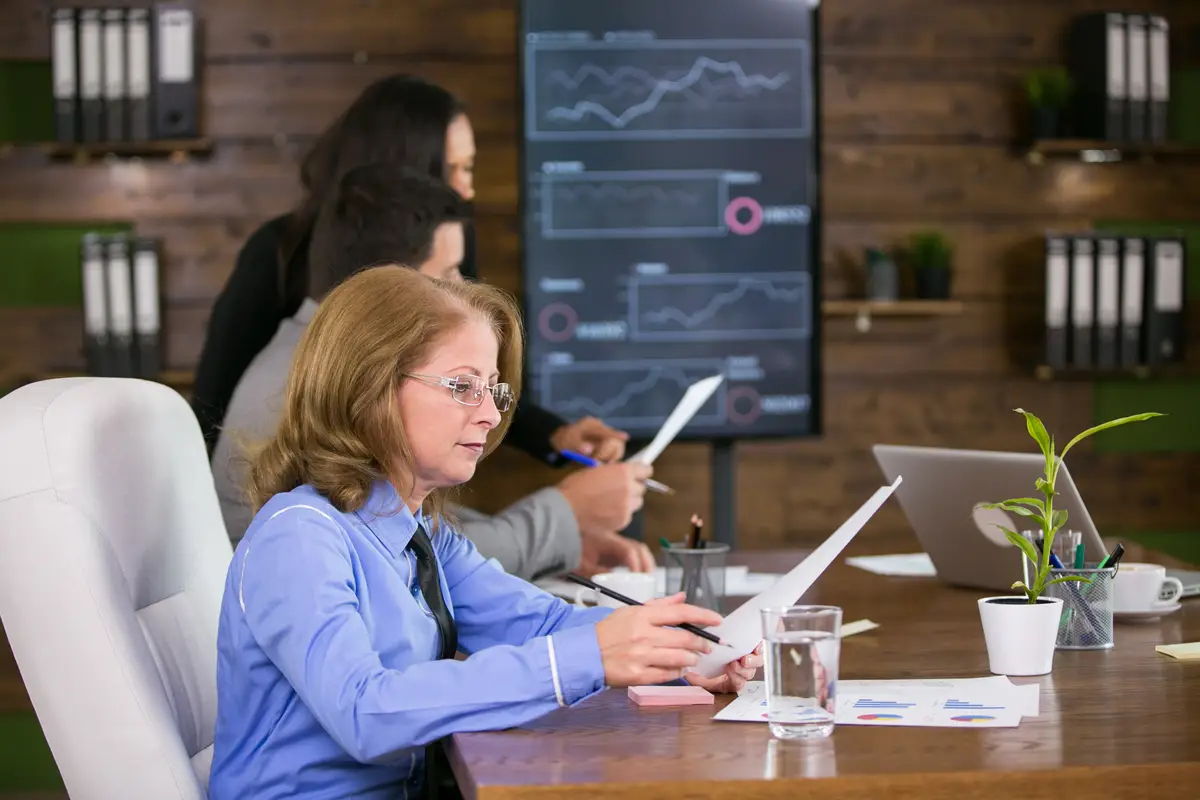 Middle age executive female reading charst in the conference room. Young businesswoman in the background.