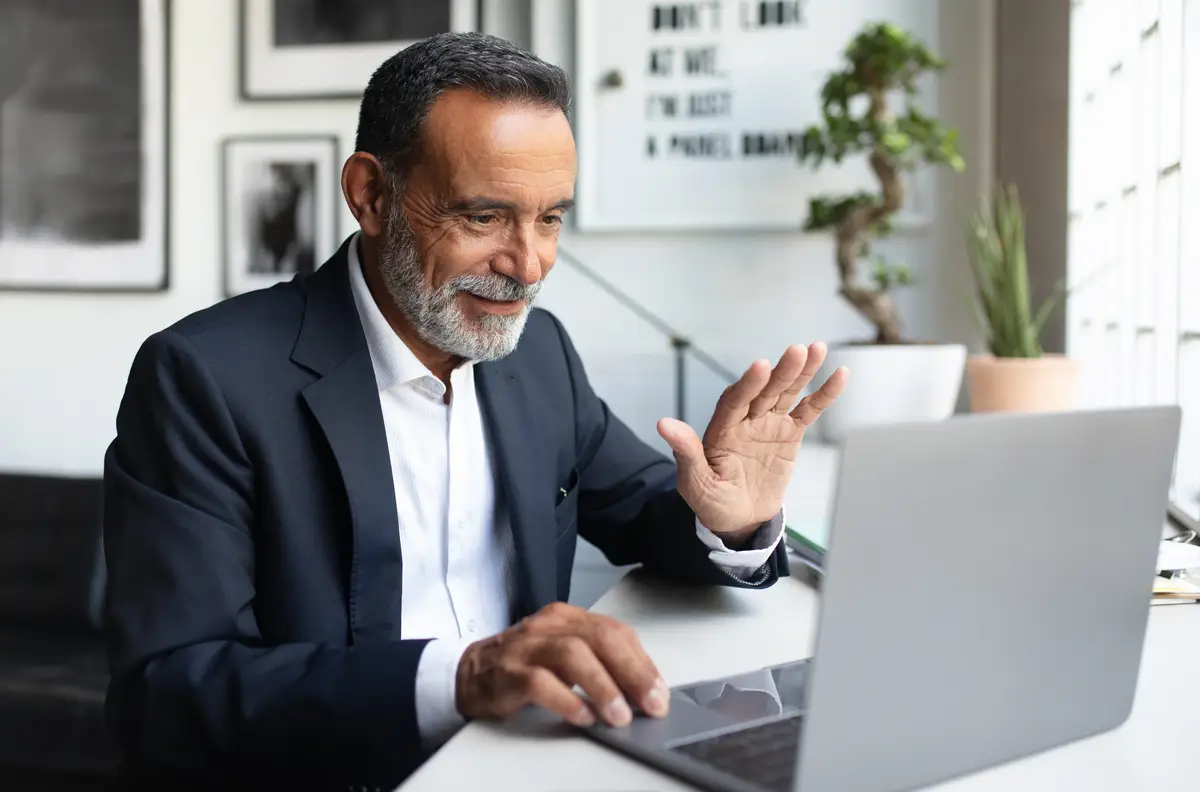 Glad mature european man waving hand say hi work on laptop in office interior greeting business