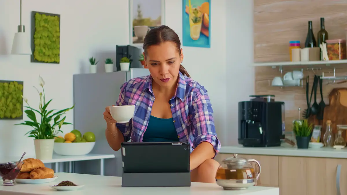 Portrait of young woman using tablet in the morning sitting at the table in the kitchen drinking tea. Working from home using device with internet technology, typing, on gadget during breakfast.