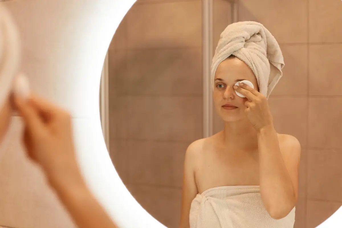 Attractive young adult Caucasian woman in towel on her hair looking in mirror and cleaning her face with cotton pad, standing in bathroom after taking shower.