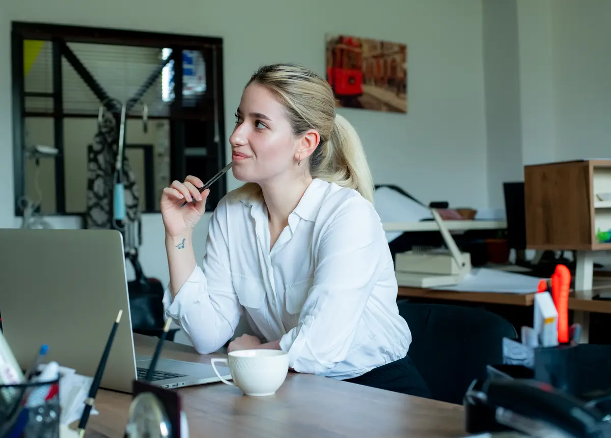 Portrait of young office worker woman sitting at office desk using laptop computer looking aside thinking with pensive expression working in office