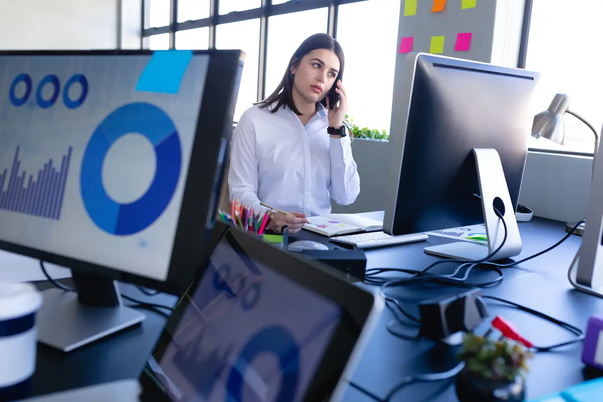 Front view of a Caucasian woman working in the modern office, sitting by a desk and using a smartphone and a desktop computer. Social distancing and self isolation in quarantine lockdown