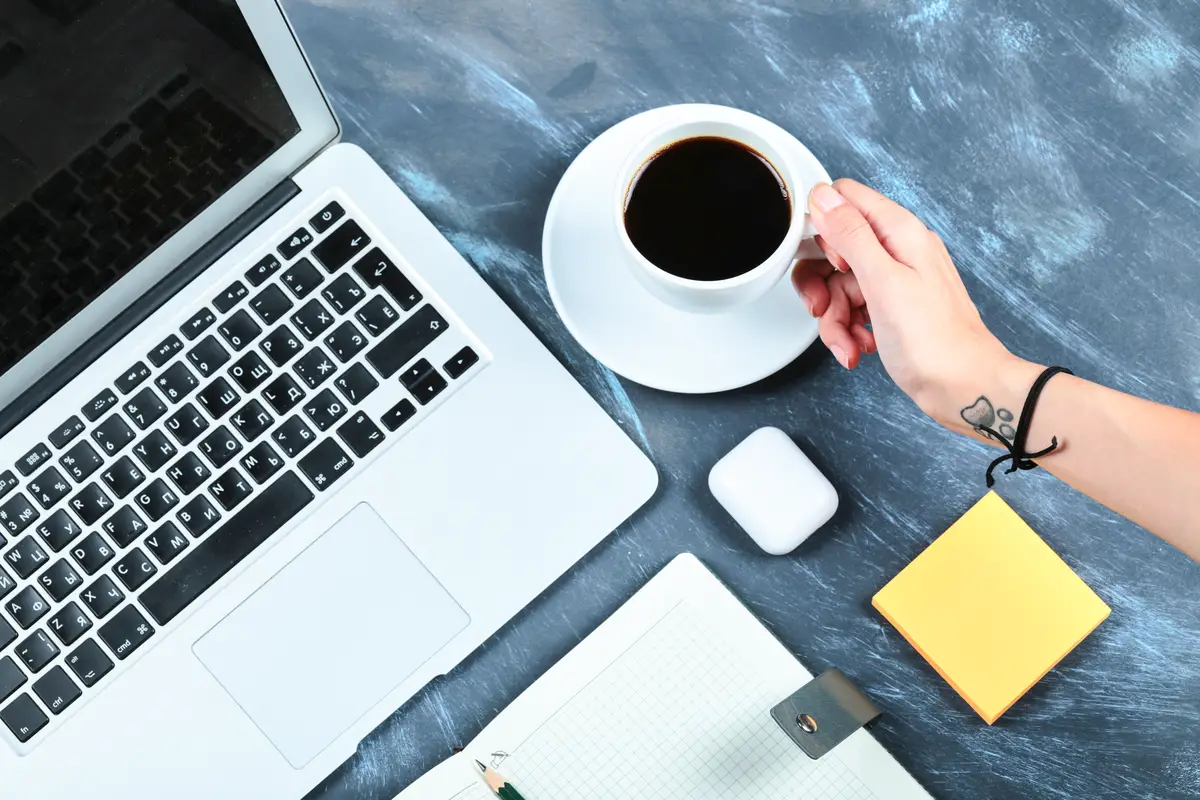 Hand holding a cup of tea with laptop, notebook, pencil and memo pads on blue desk.