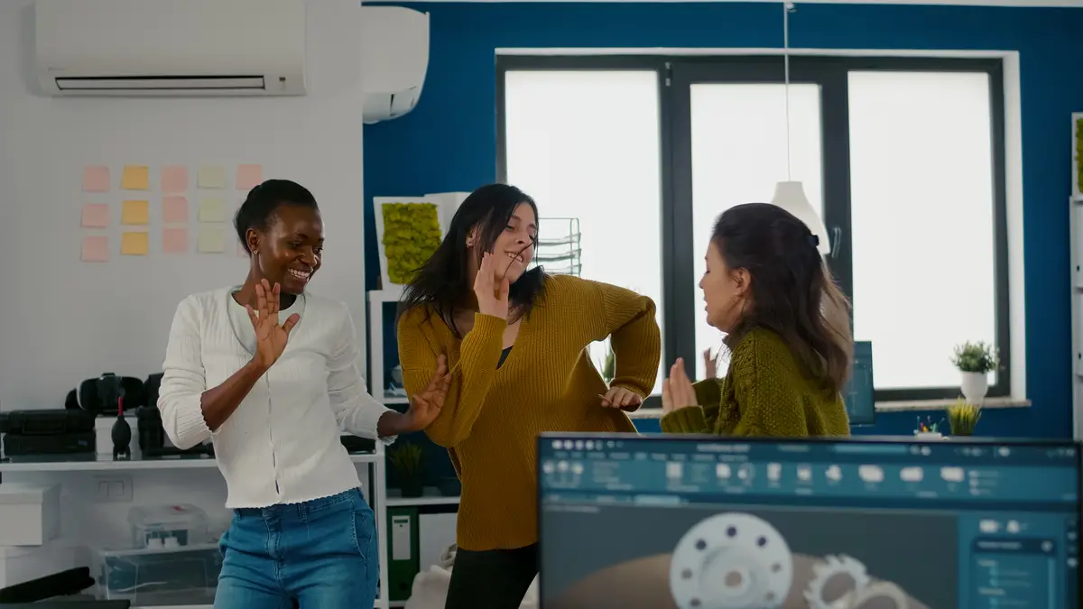 Enthusiastic diverse engineers women dancing in company office celebrating success of project singin...