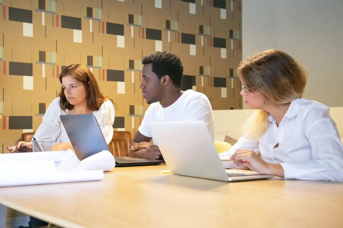 Diverse group of designers working on project together, sitting at table with laptops and blueprints and talking