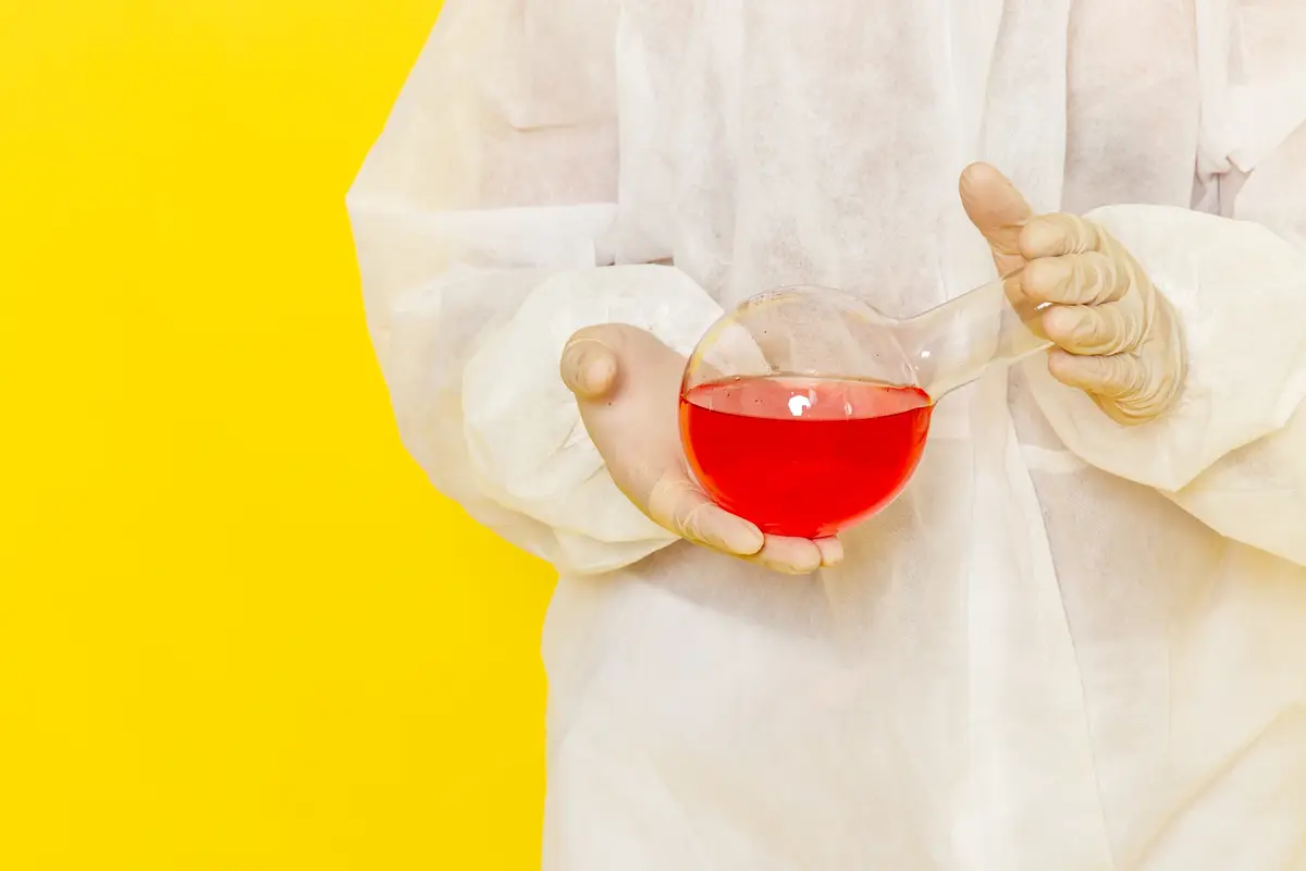 Front close view male scientific worker in special protective suit holding flask with red solution on light yellow desk