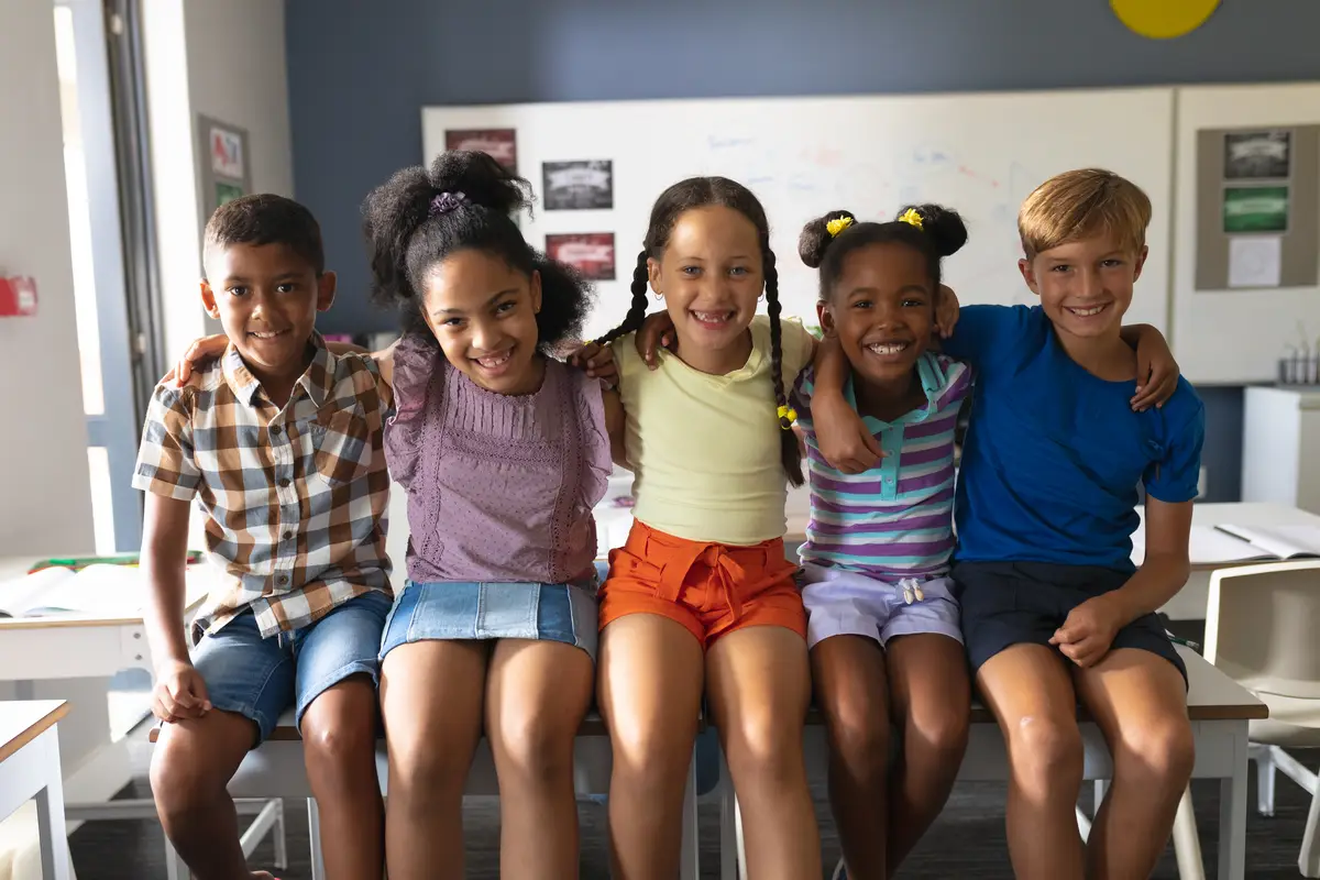 Portrait of smiling multiracial elementary school students sitting with arm around in classroom