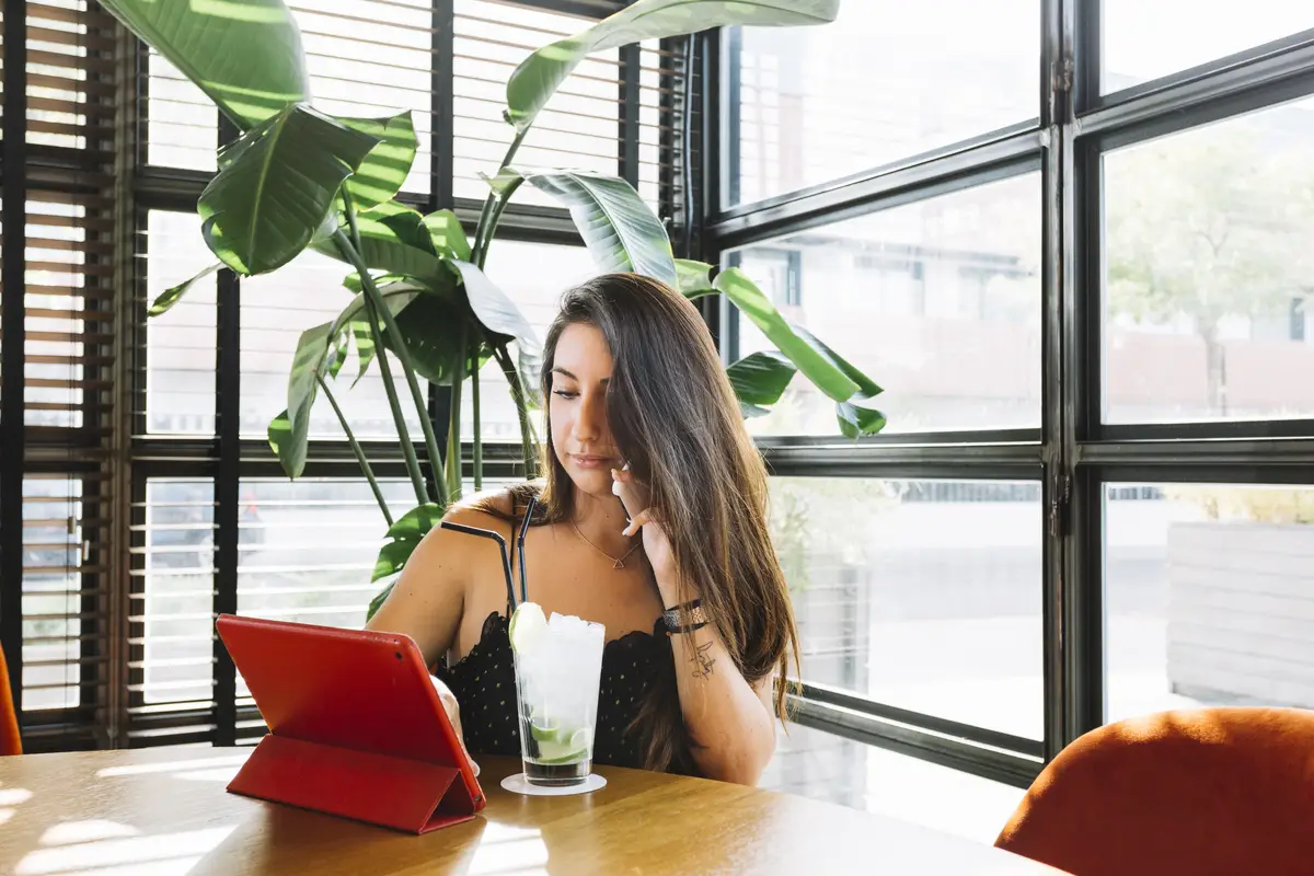 Young woman talking on cellphone using digital tablet with cocktail on table