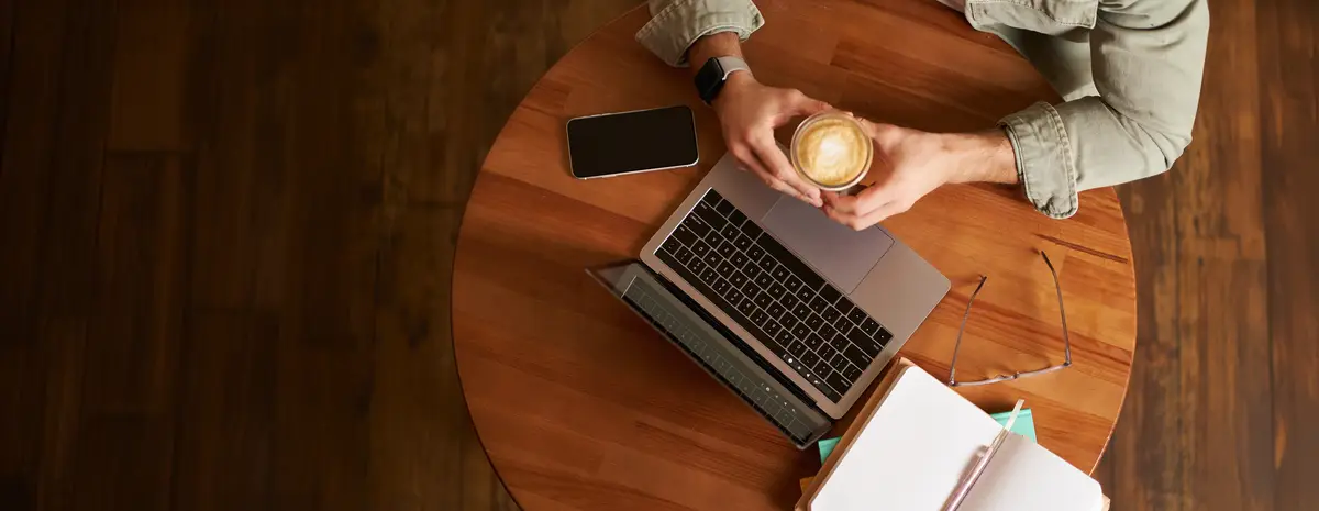 Top view Male hands holding cup of coffee man sitting at round table in a cafe working on laptop had his smartphone and notebook drinking cappuccino