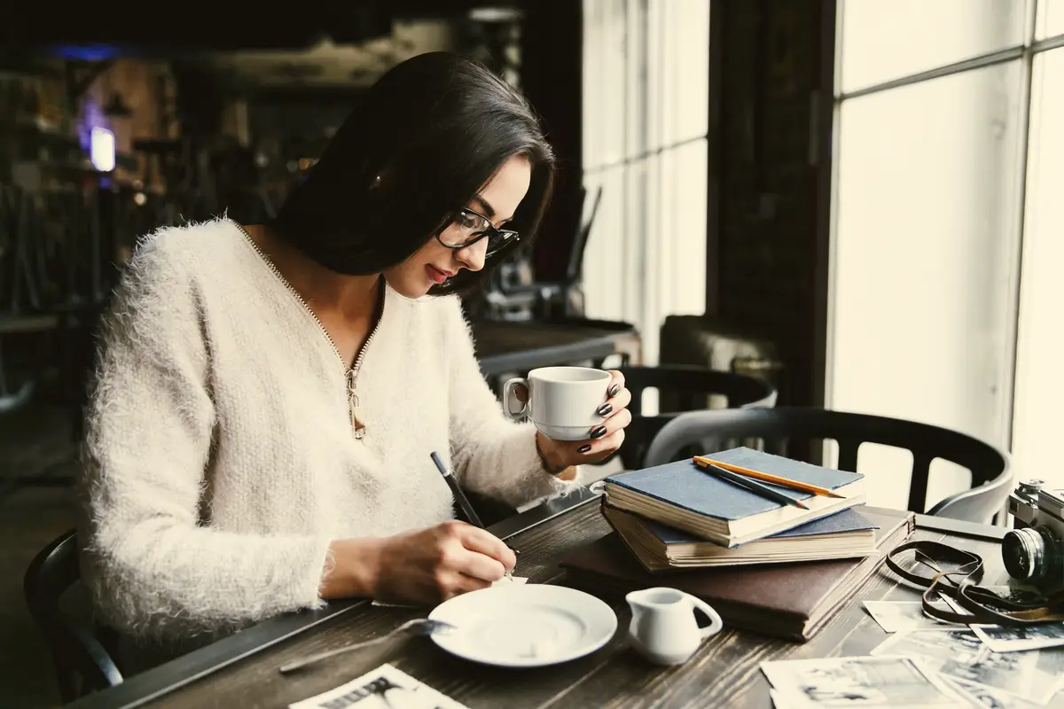 Girl with long hair drinks coffee at the table in cafe and makes some notes