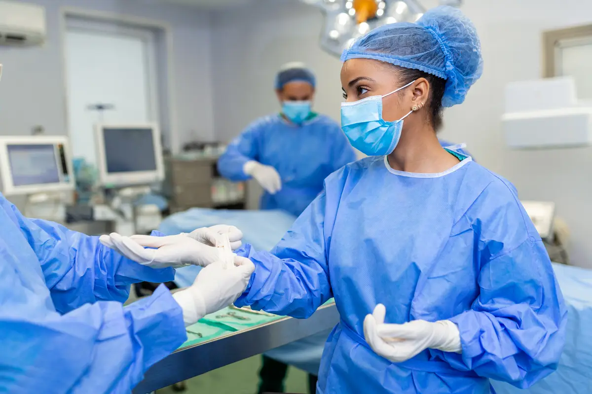 Indicações cirúrgicas na endocardite: recomendações atuais 3 Young nurse in protective mask and workwear helping surgeon with gloves while both preparing for operation