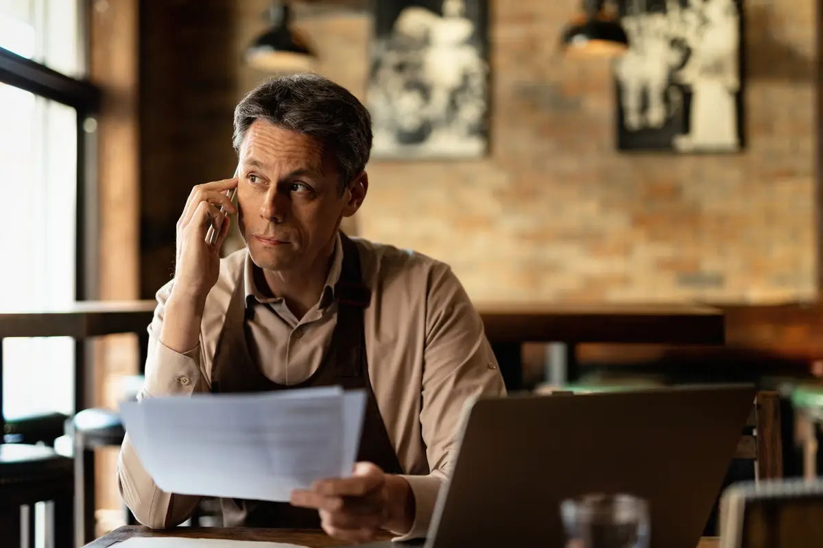 Mid adult bar owner analyzing documents and talking on mobile phone after working hours