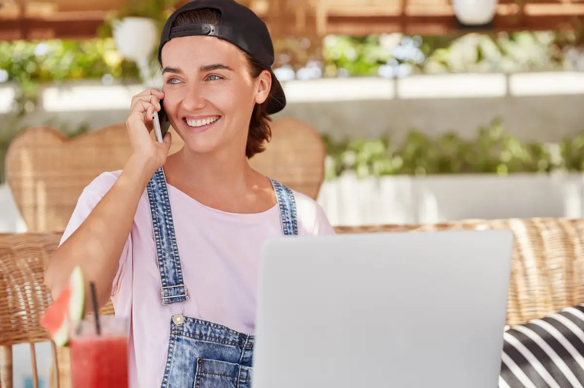 Photo of glad blue eyed woman in fashionable cap, speaks with friend on smart phone, looks happily away, downloads news application on portable laptop computer, drinks fresh cocktail in cafe