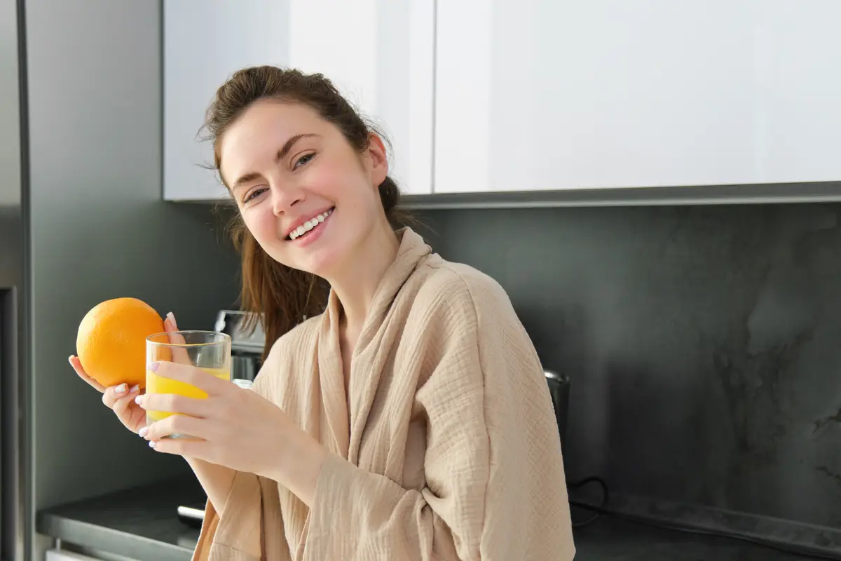 Beautiful young woman in bathrobe drinking homemade orange juice smiling and laughing standing near