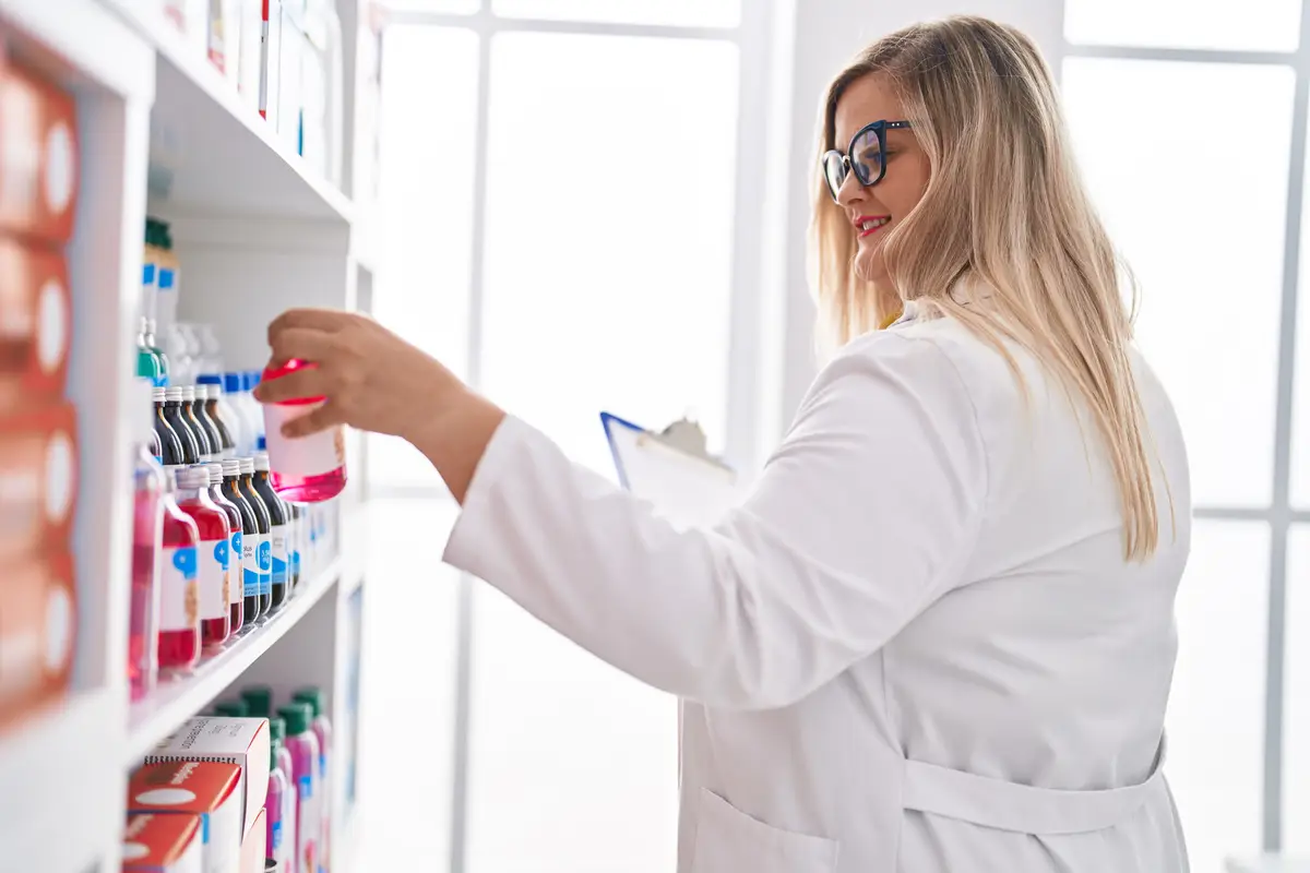 Young woman pharmacist reading document holding medication bottle at pharmacy