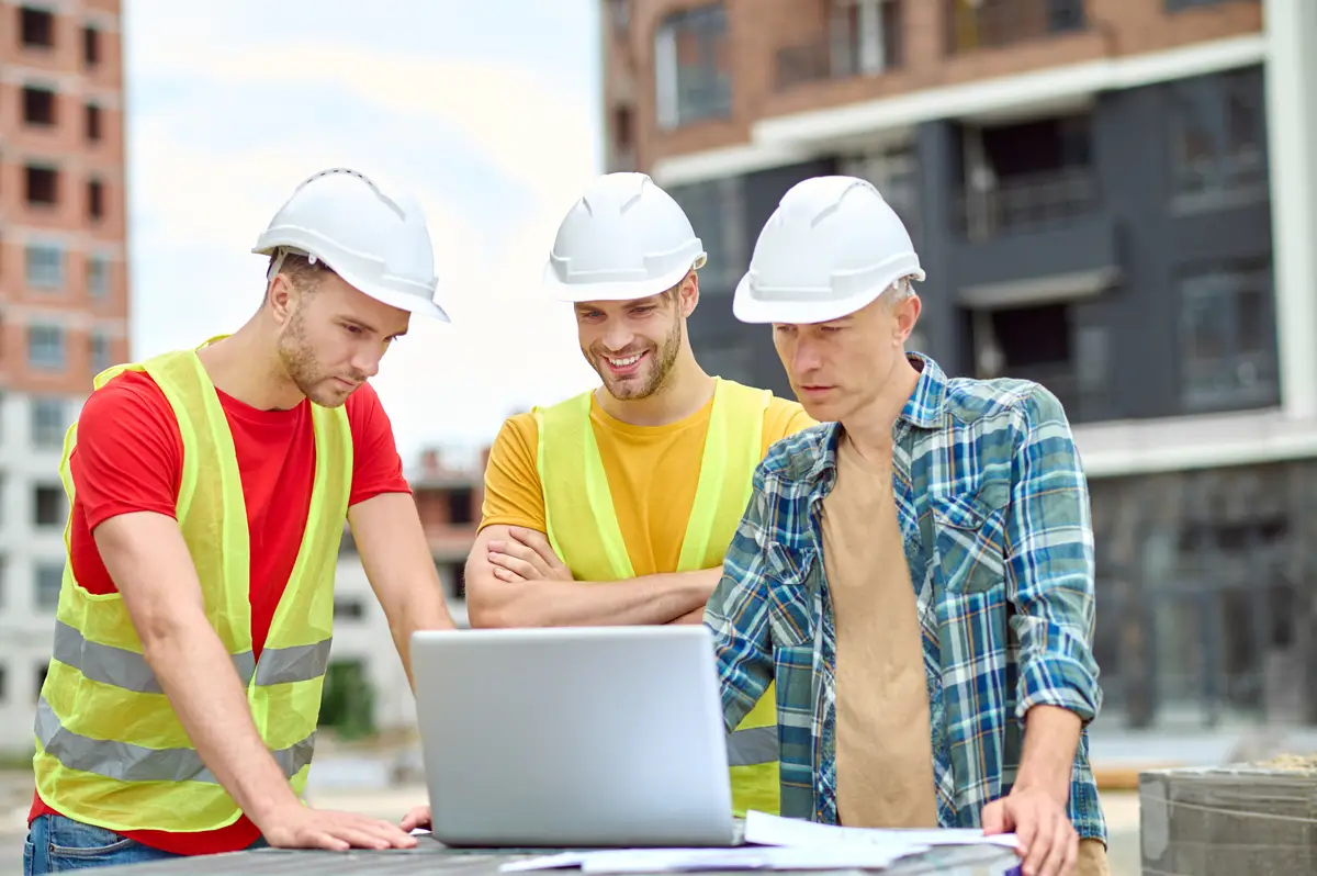 Important information. Three men in protective helmet looking at laptop with interest while standing at construction site during day