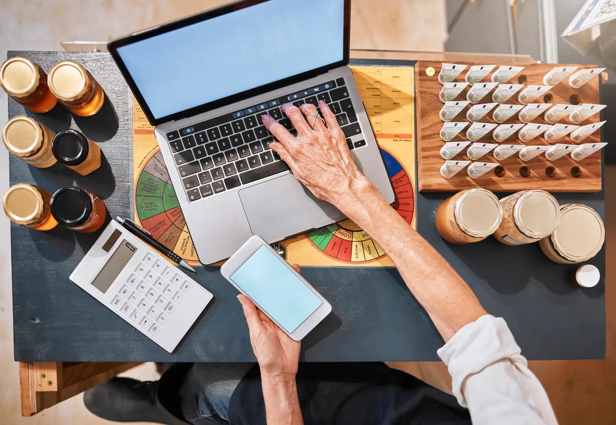 Hands laptop and honey with a retail woman checking her phone while working at a desk in her store Computer ecommerce and email with a female employee typing a text message at work in her startup