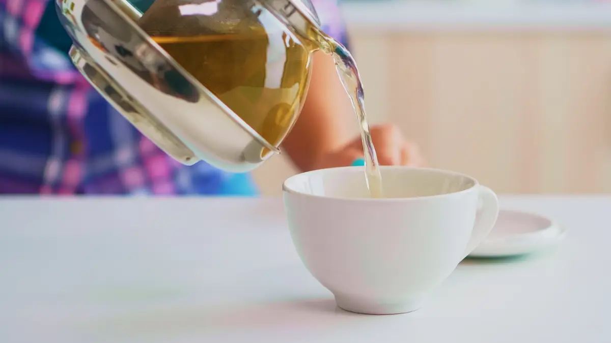 Green tea flowing from teapot in slow motion. Close up of tea from the kettle slowly pour into porcelain cup in the kitchen in the morning at breakfast, using teacup and healty herbal leaves.