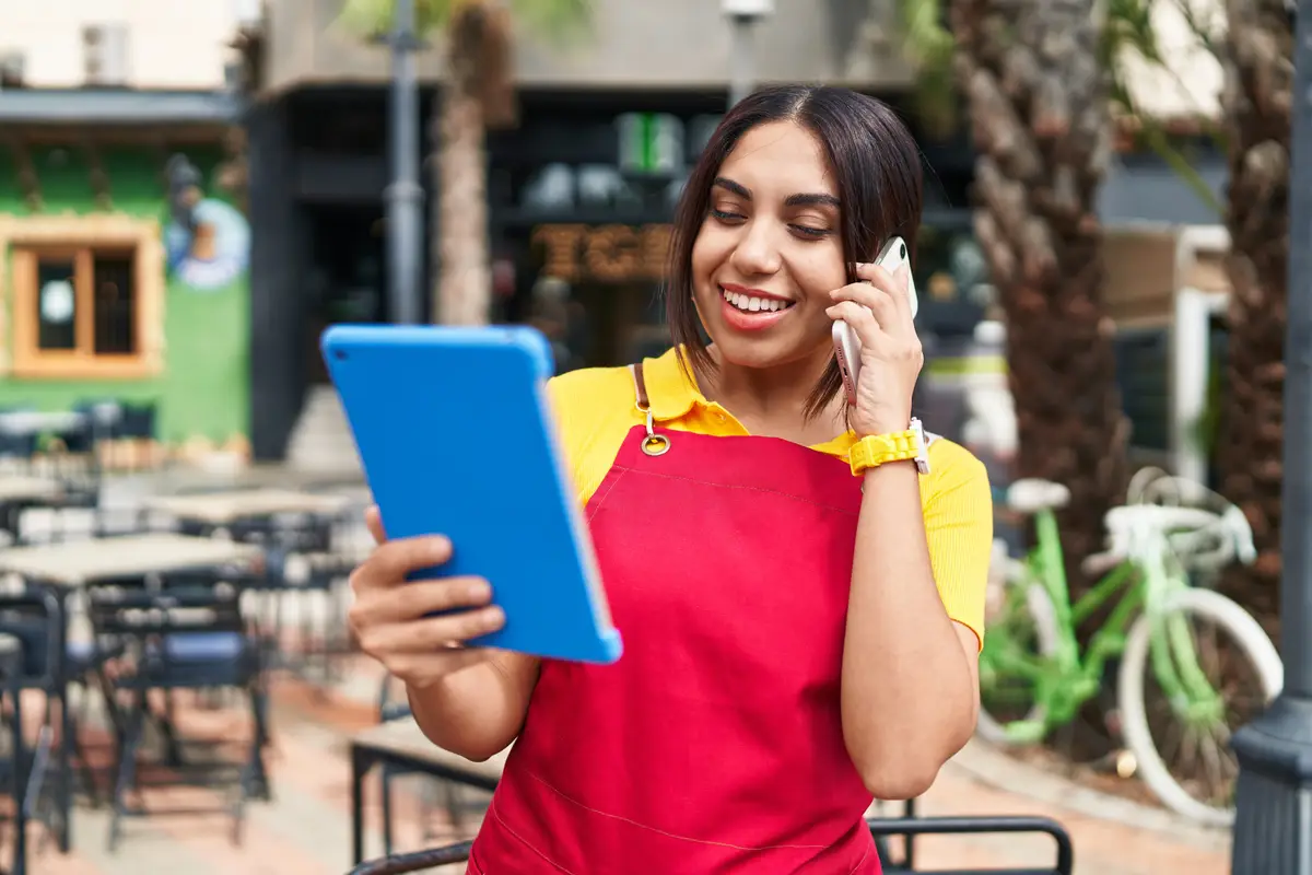 Young beautiful arab woman waitress talking on smartphone using touchpad at coffee shop terrace